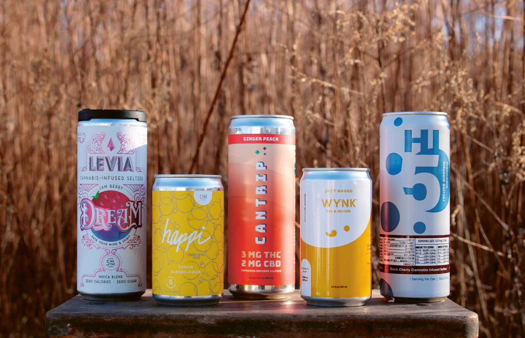 A lineup of 5 cannabis-infused seltzer cans in front of a backdrop of dried tall grass.