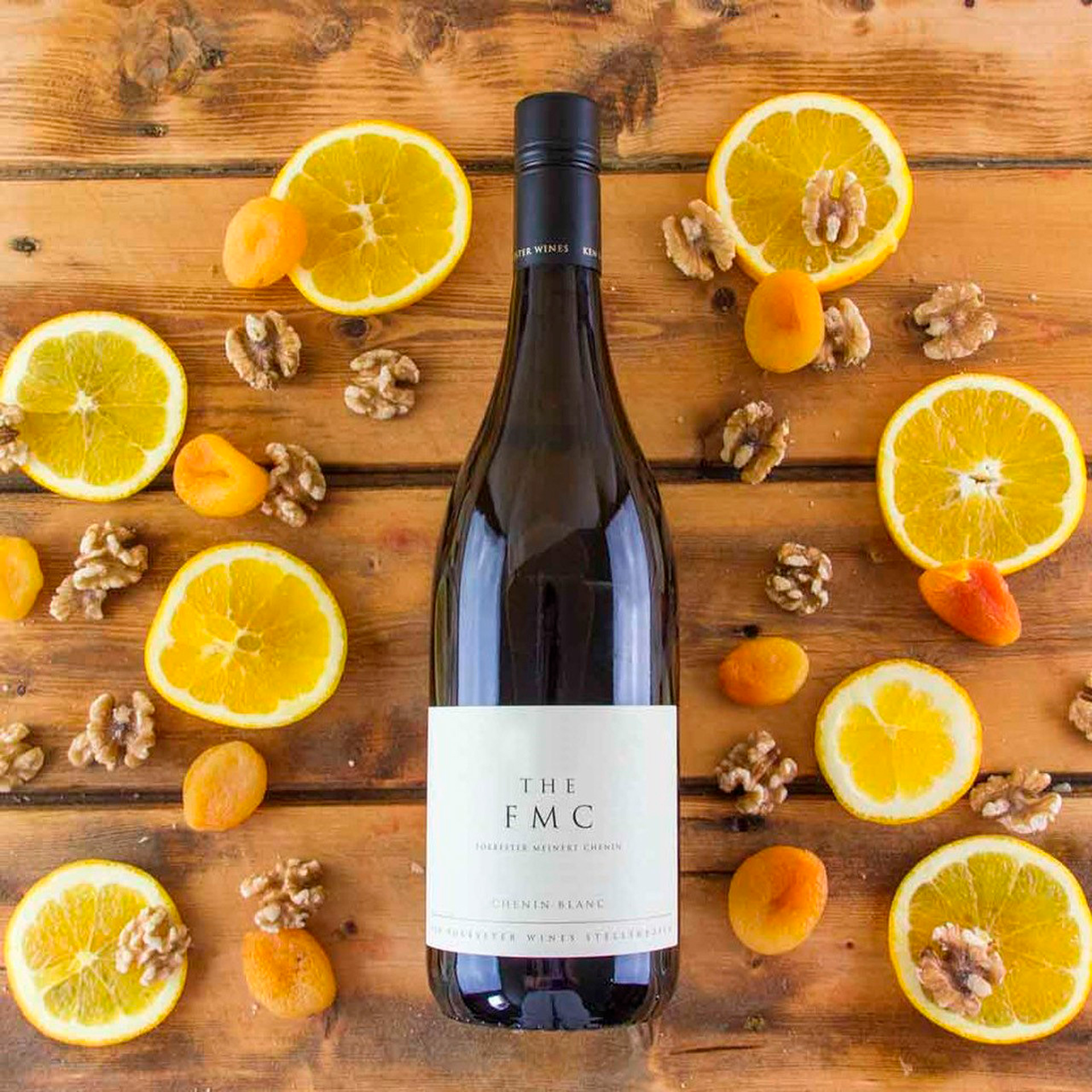 Image of a bottle of Ken Forrester Wines FMC lying on a table, surrounded by descriptors of the wine's profile, including lemons, oranges, apricots and walnuts.