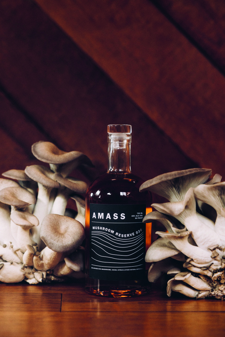 A bottle of Amass Mushroom Gin on a wood table with mushrooms surrounding it.