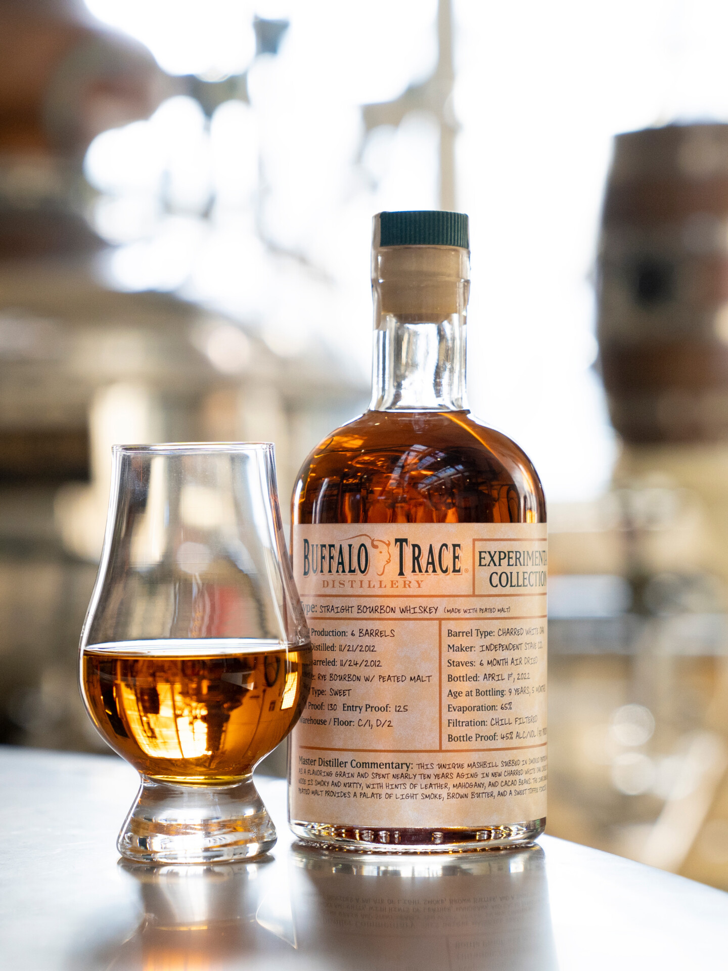 A bottle from Buffalo Trace Distillery with a poured tasting glass next to it, with barrels and a kiln int he background.