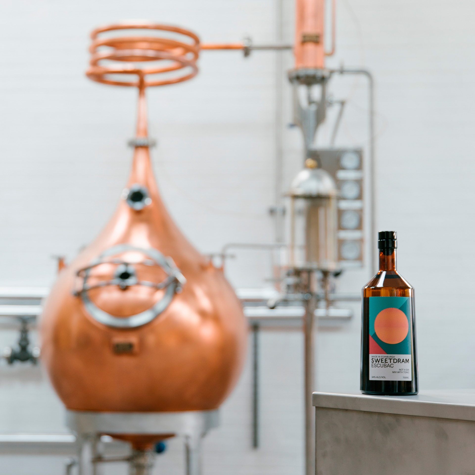 A bottle of Sweetdram Escubac on a table in the foreground, with a copper distilling kiln in the background.