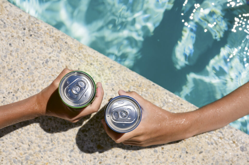 Two hands, each holding an aluminum can in hand, resting on a poolside surface.