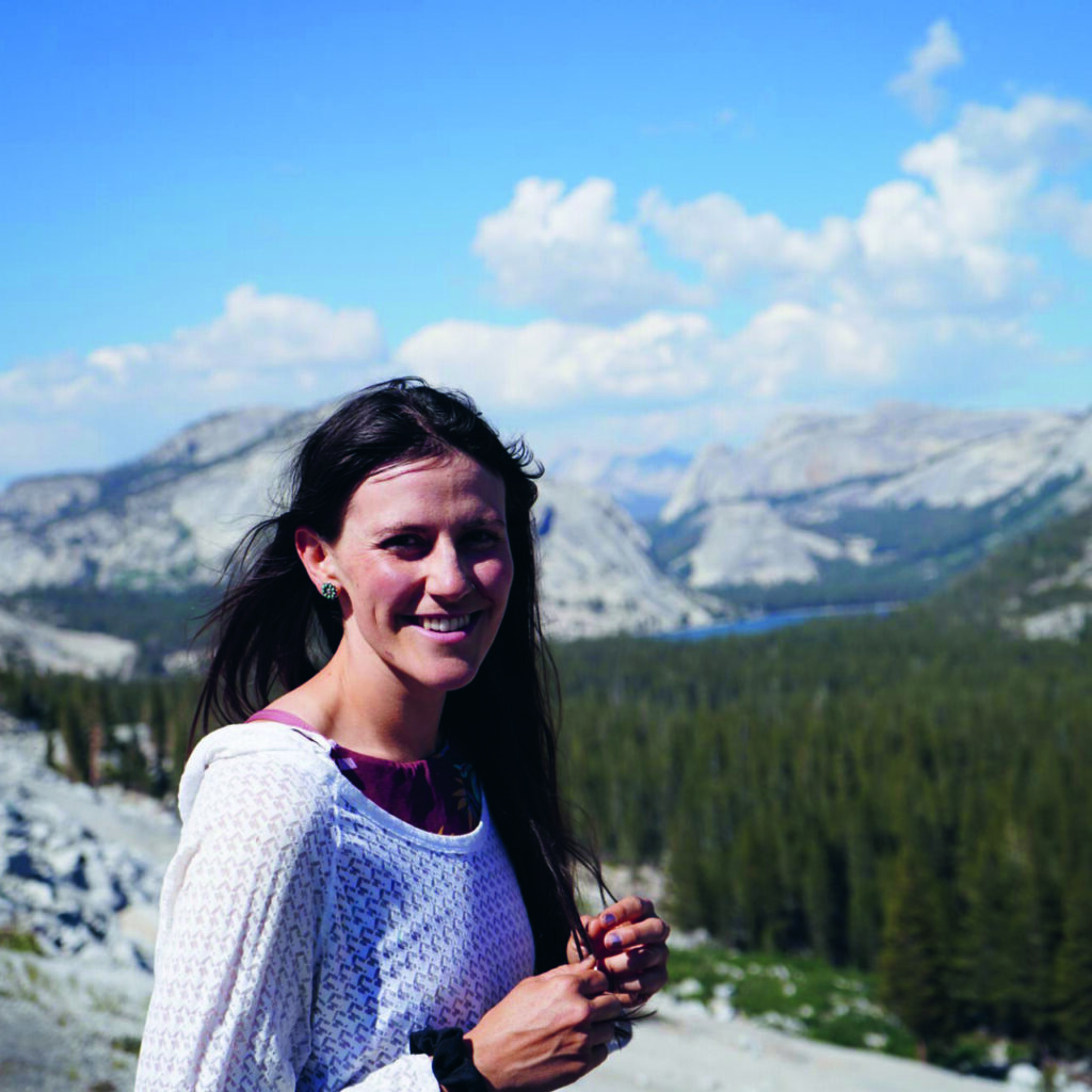 Picture of writer Helena Guglielmino, sitting atop a mountain with mountains in background and a glass in hand.