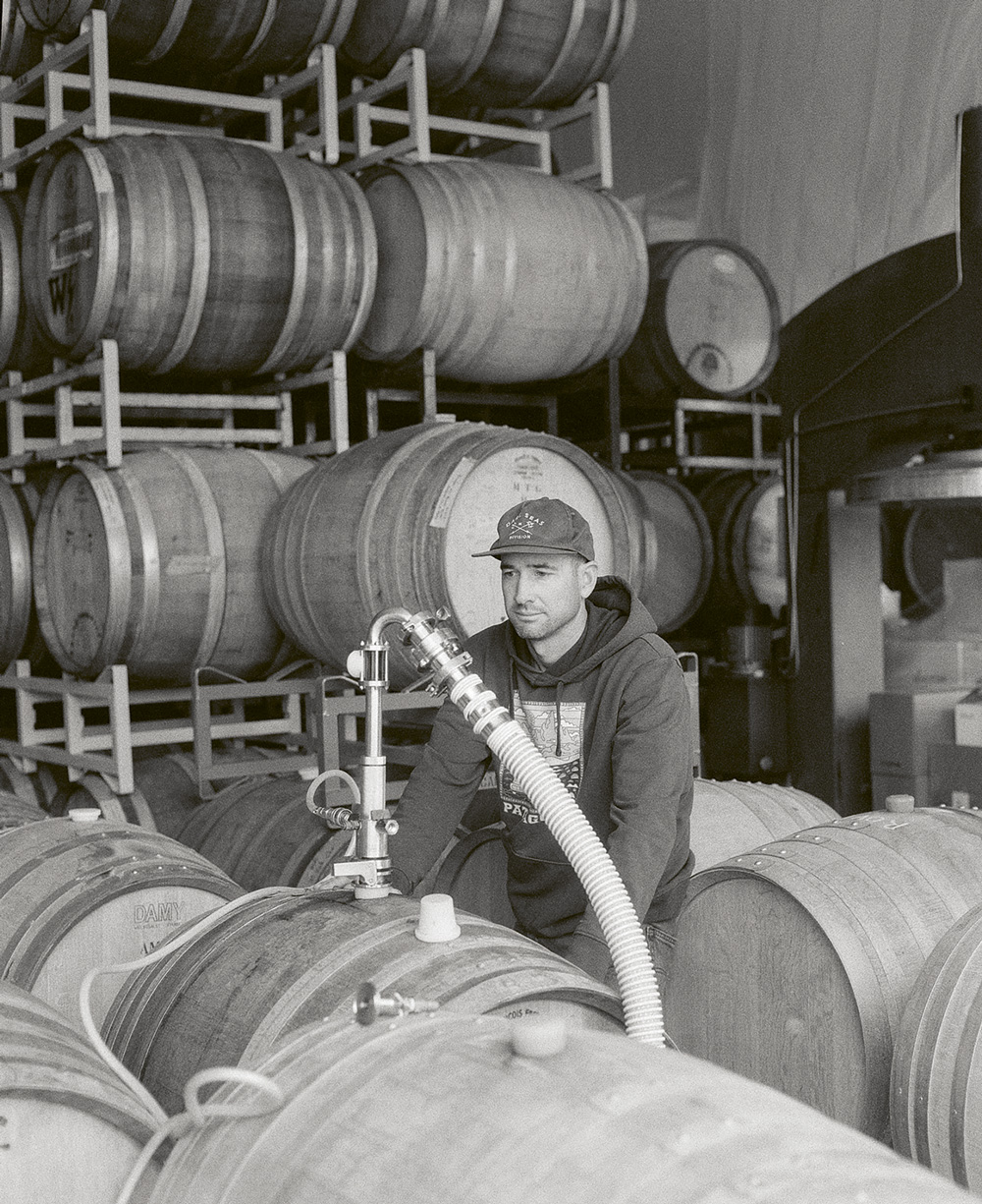 A black and white photograph of winemaker Mikey Giugni of Scar of the Sea seen standing surrounded by wood barrels with a hose in one, filling it.