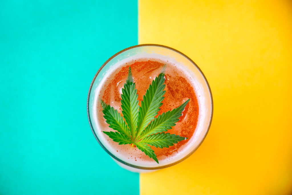 Overhead picture of a glass with liquid and ice and a cannabis leaf floating atop, set on a background of teal and yellow.