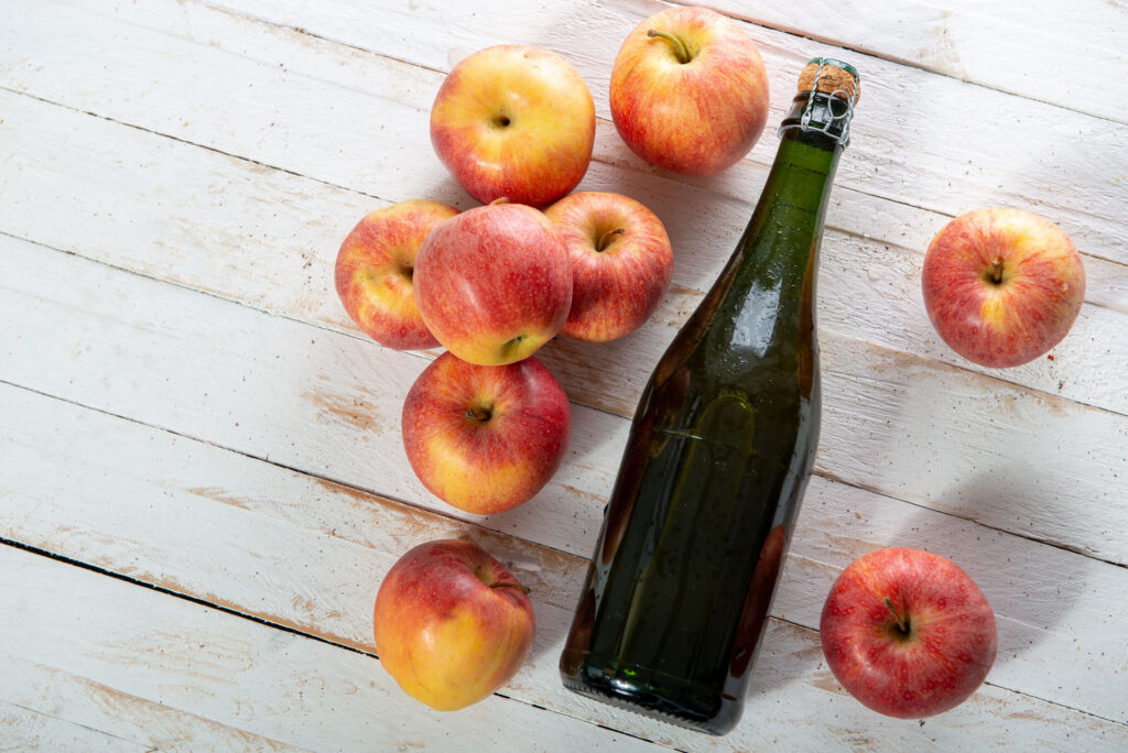 cider-hero A cork-topped unlabeled bottle resting on a white wood background with an assortment of yellow-red apples alongside it.