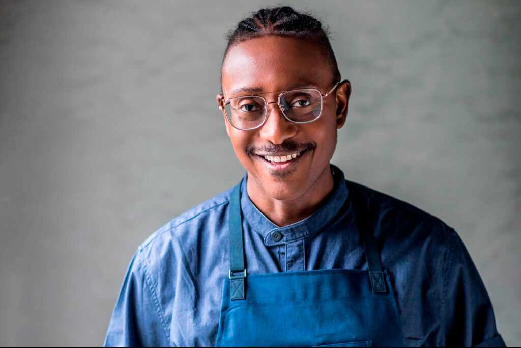 A headshot of Chef Gregory Gourdet, wearing a blue shirt and blue apron.