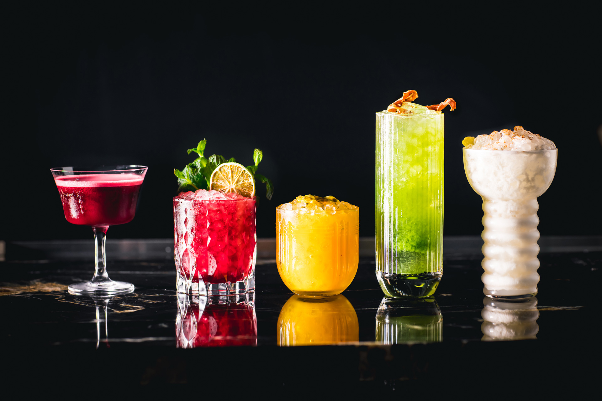 A selection of five mixed drinks lined up on a black counter, shot head on, in assorted colors and glasses, made at Sousol in Portland, Oregon.