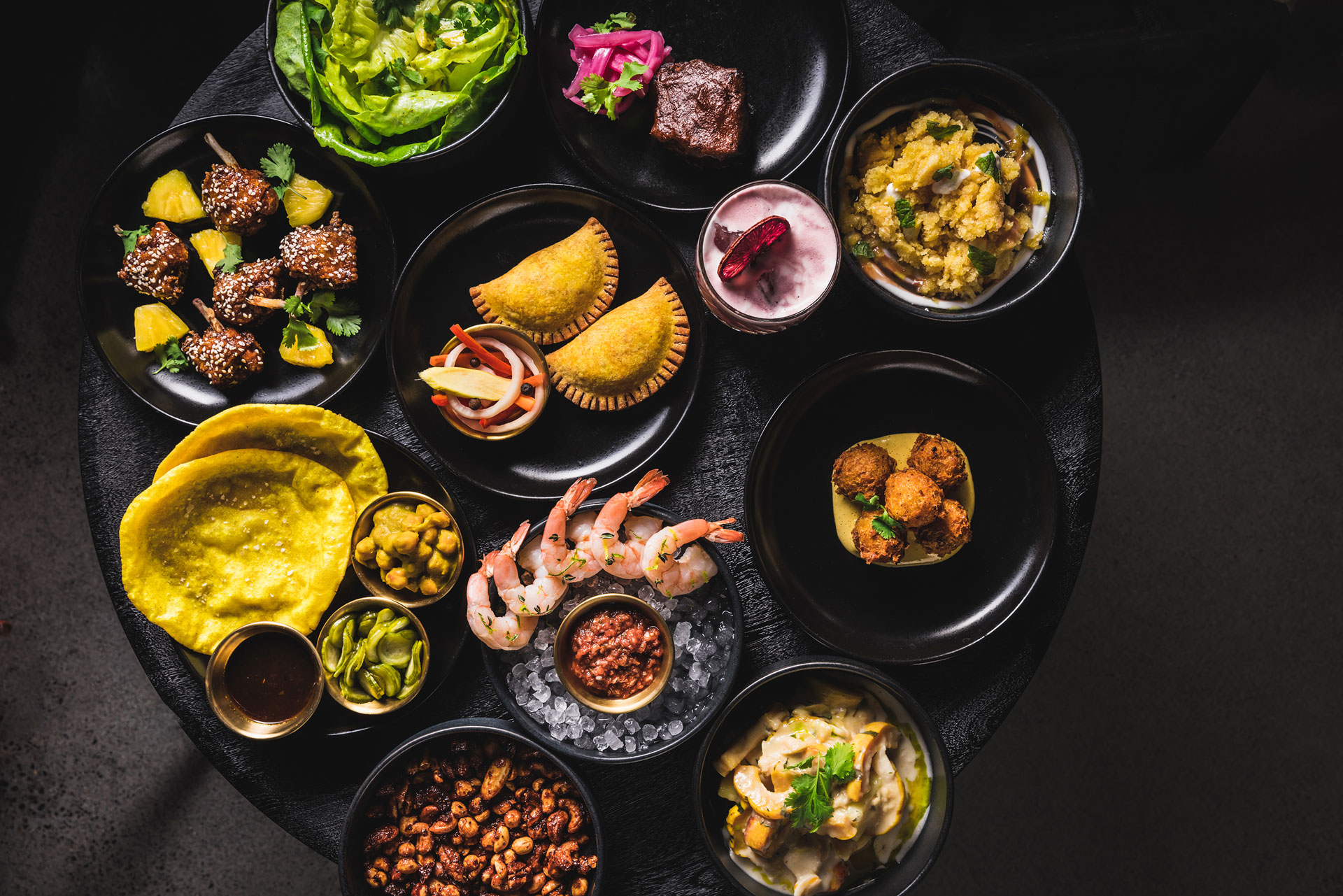 A selection of foods prepared at Sousol in Portland, Oregon, including empanadas, shrimp cocktail, braised pork belly and fritters, all plated on black dishes and on a black tabletop, shot overhead.