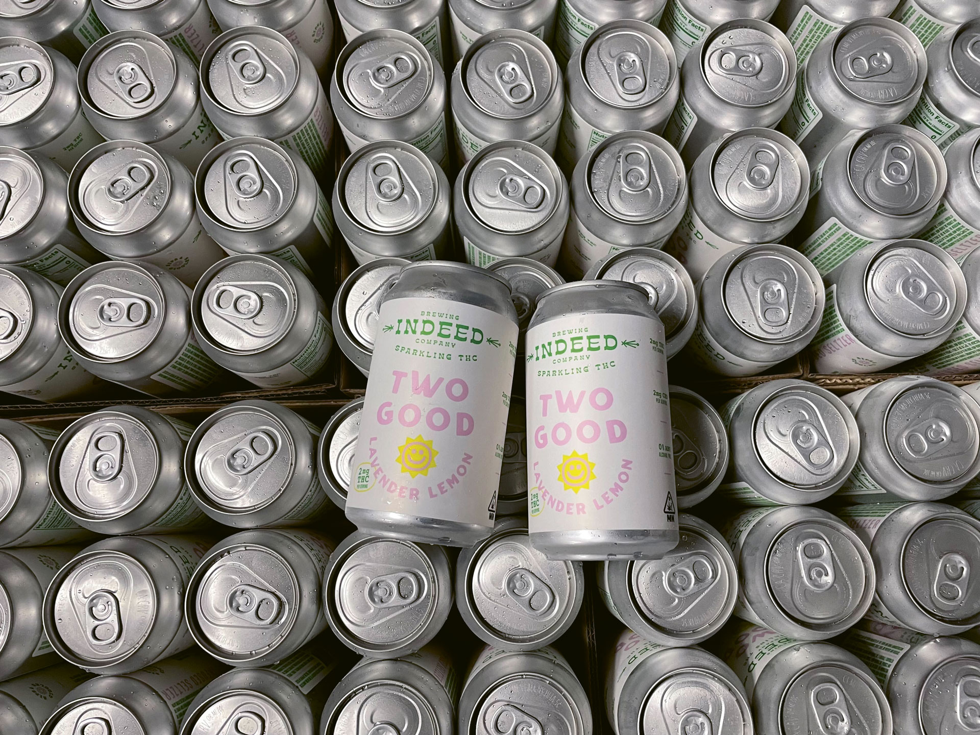 Two horizontal cans of Two Good from Indeed Brewing atop a stack of upright rans.