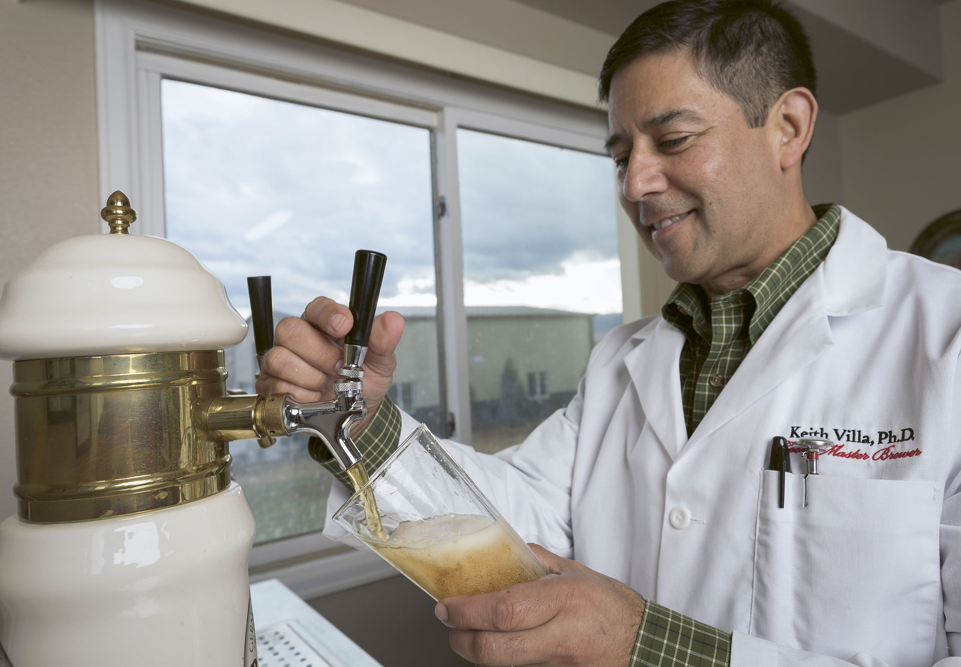 Keith Villa, founder and brewmaster of Ceria Brewing, in a labcoat pulling a draught sample from a tabletop keg.