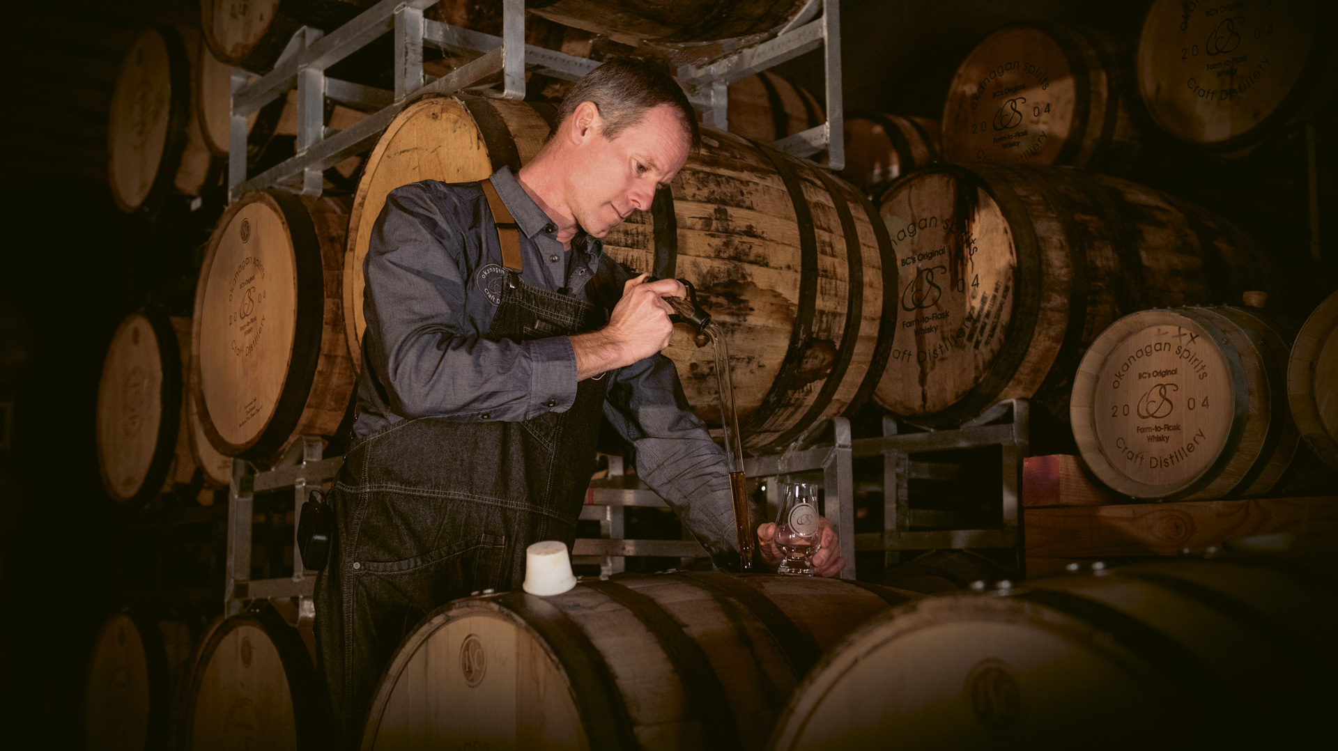 Tyler Dyck of Okanagan Spirits in pictured surrounded by barrels with a thief in hand, which is filling up a tastin glass in the other hand.