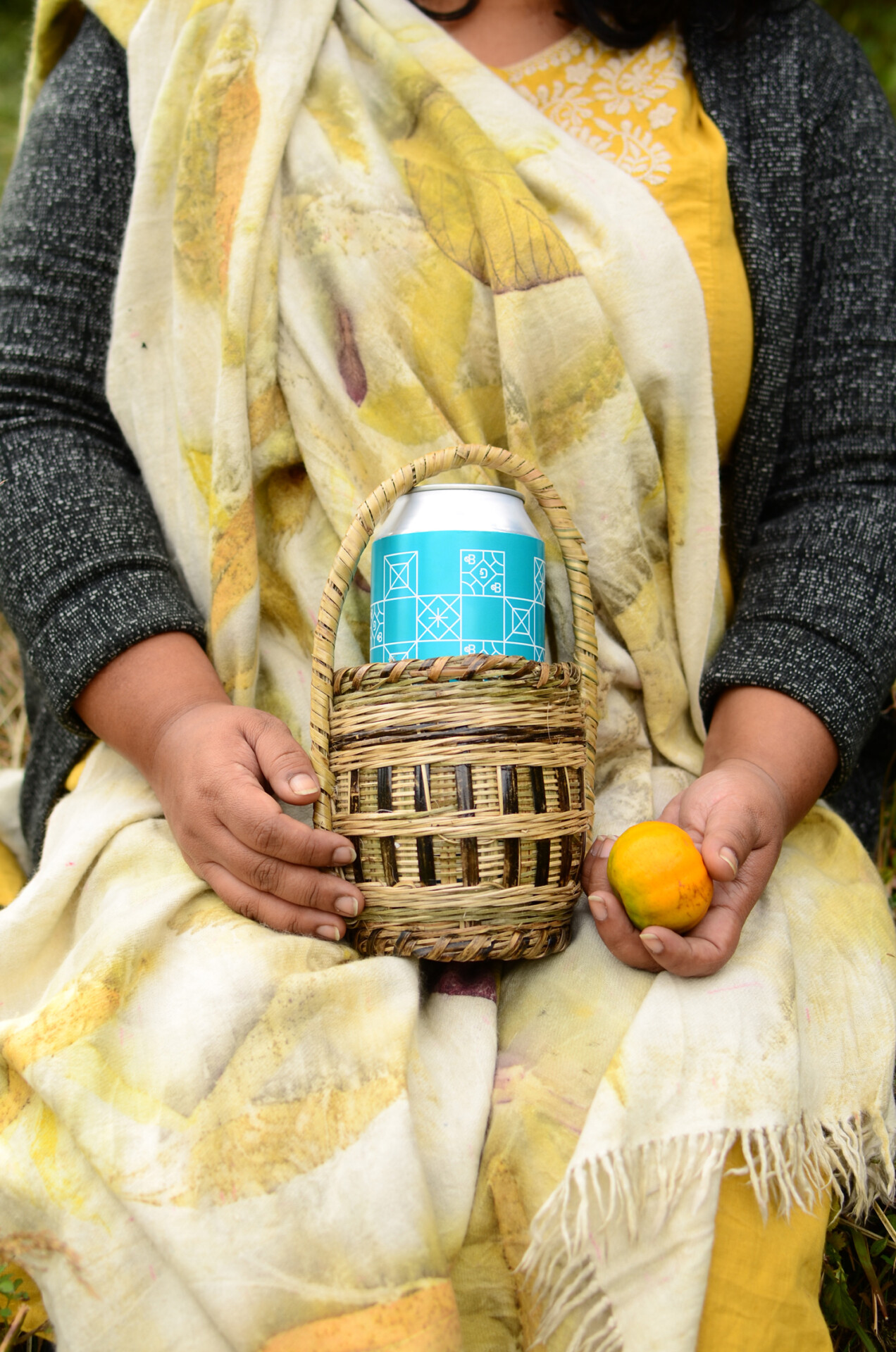 A seated woman holds a basket with a can of Bombay Duck Brewing beer in one hand and a locally sourced citrus fruit in the other