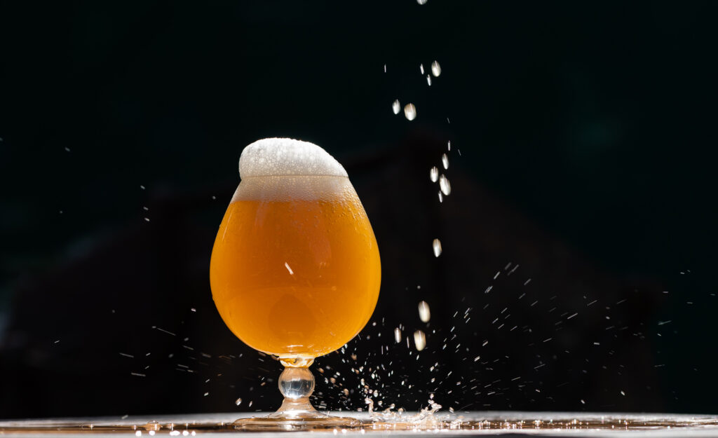 A full golden tulip of beer set on a table against a black backdrop, with drops falling from overhead.