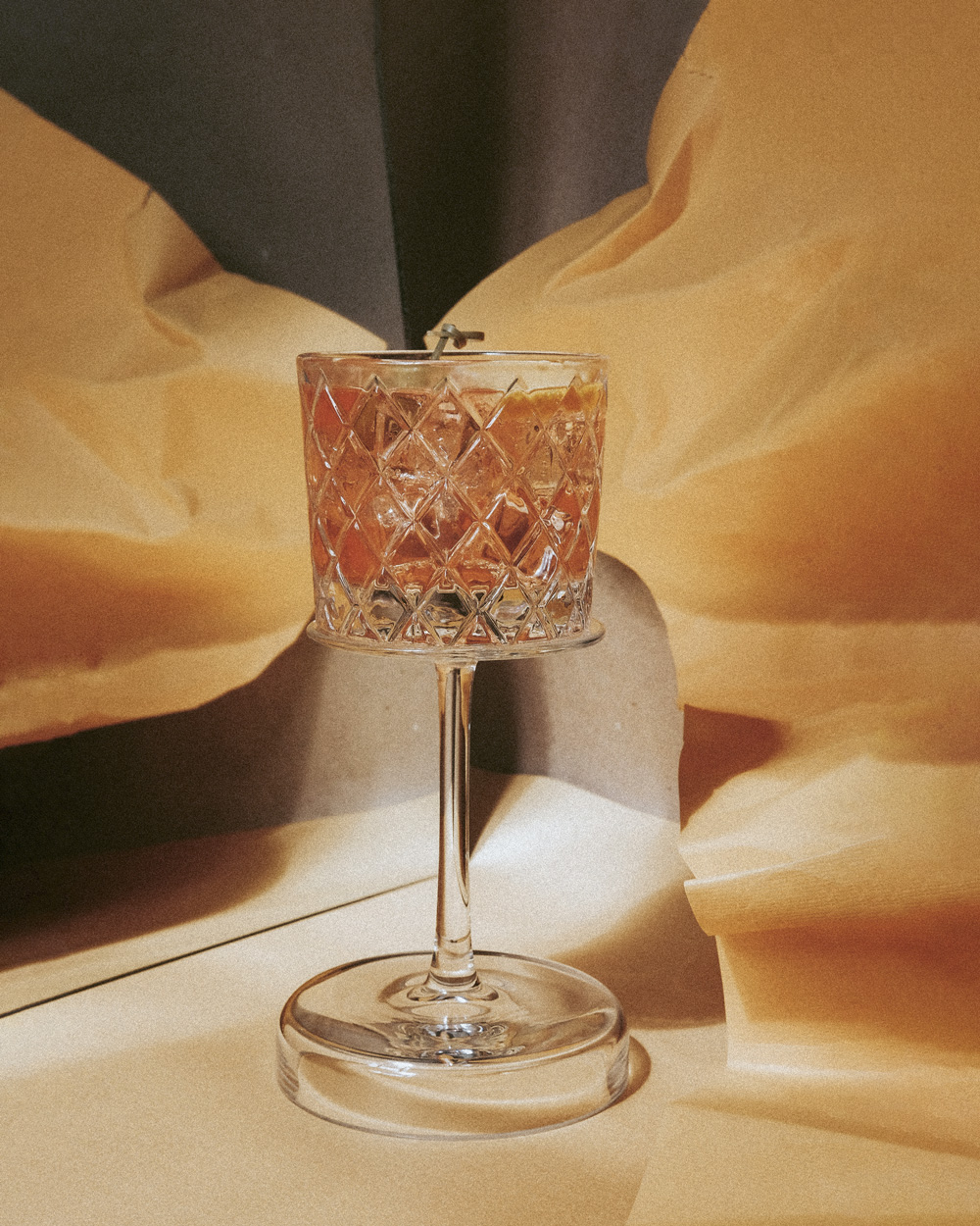 A stemmed drink glass with a rose-hueded cocktail in it and gold-colored fabric in background.
