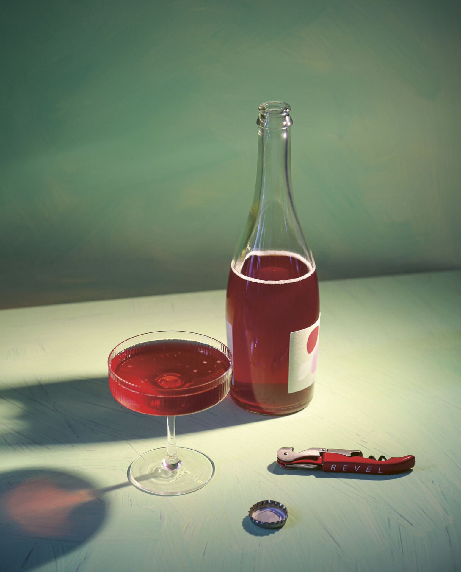 A bottle of Revel Cider Soif set atop a green table with a green background, standing alongside a poured glass with long shadows.