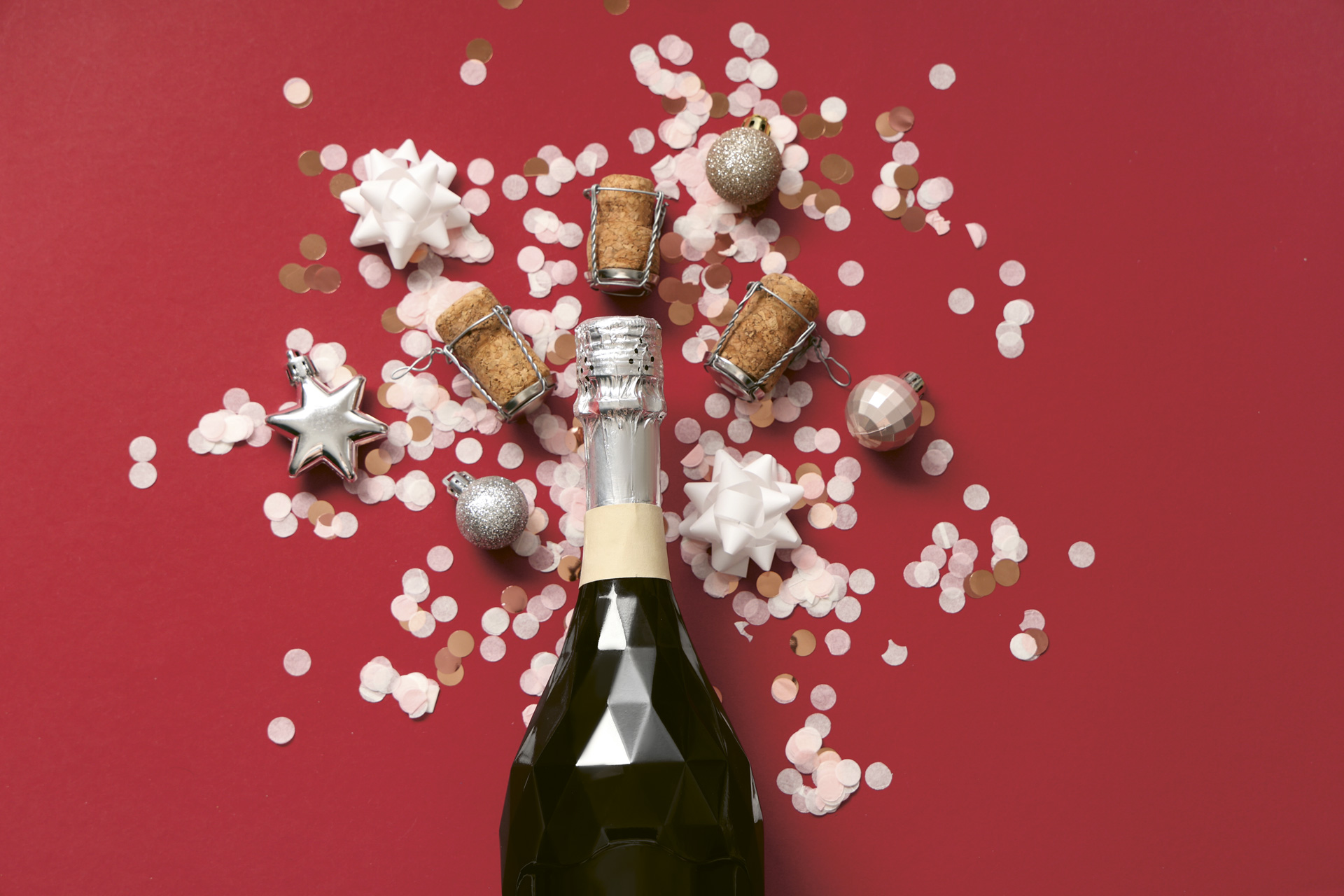 A picture of a Champagne bottle on a red background with corks and holiday confetti surrounding it.
