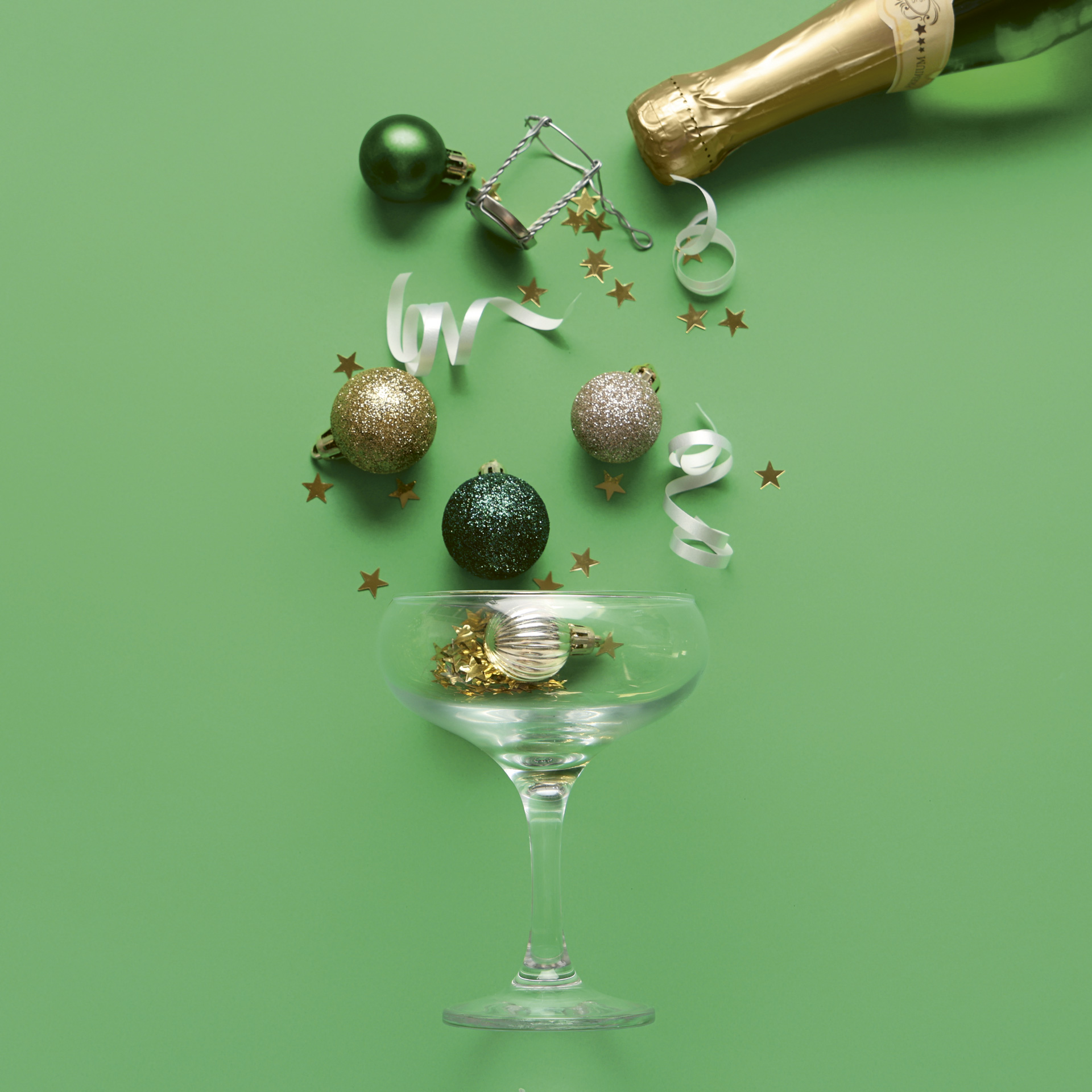 A picture of a Champagne coupe on a green background with ornaments and holiday confetti pouring into it from a sparkling wine bottle at top right.
