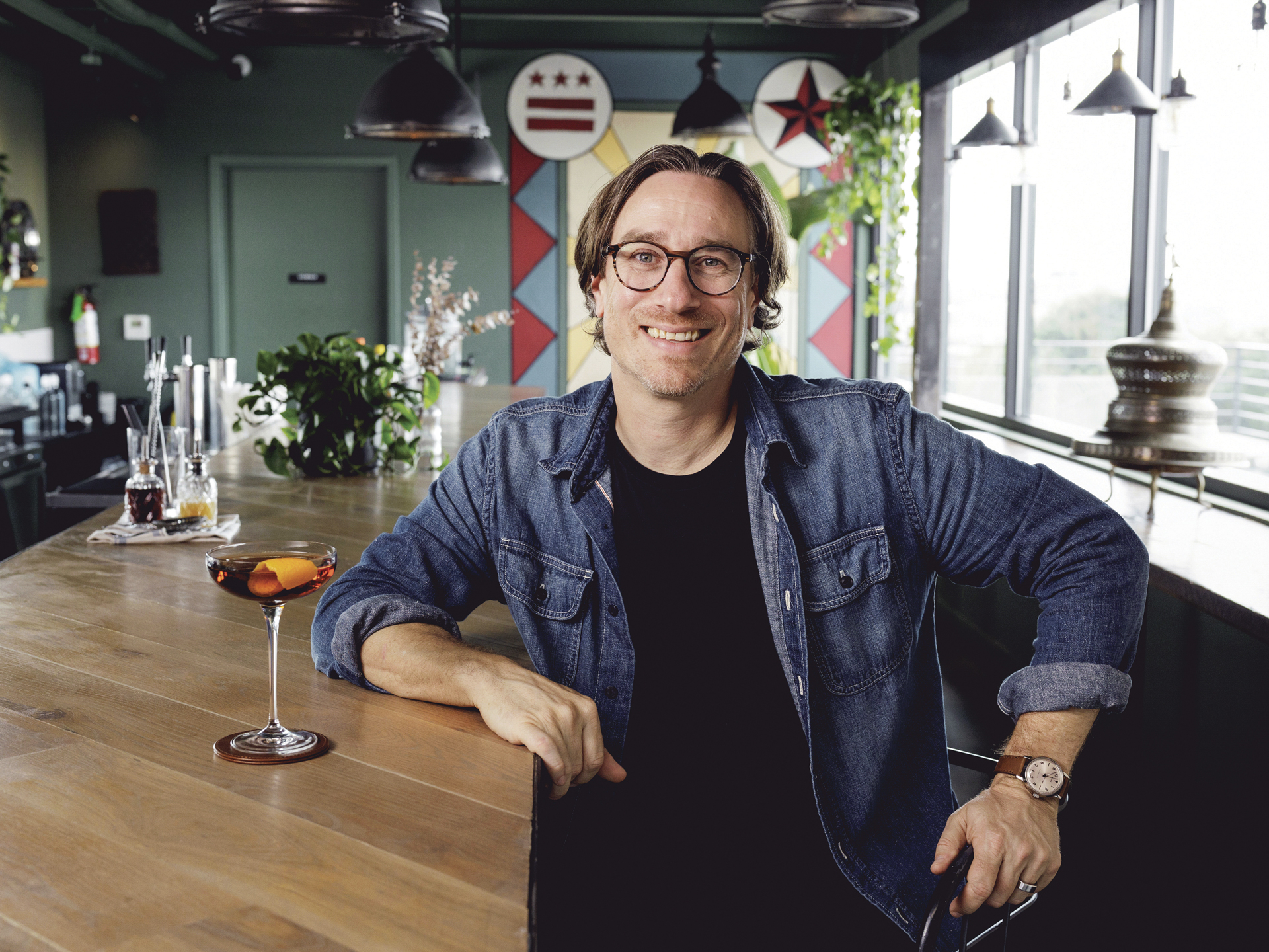 Mixologist and author Derek Brown sitting at a bar smiling, with a couped cocktail on the bar in front of him.