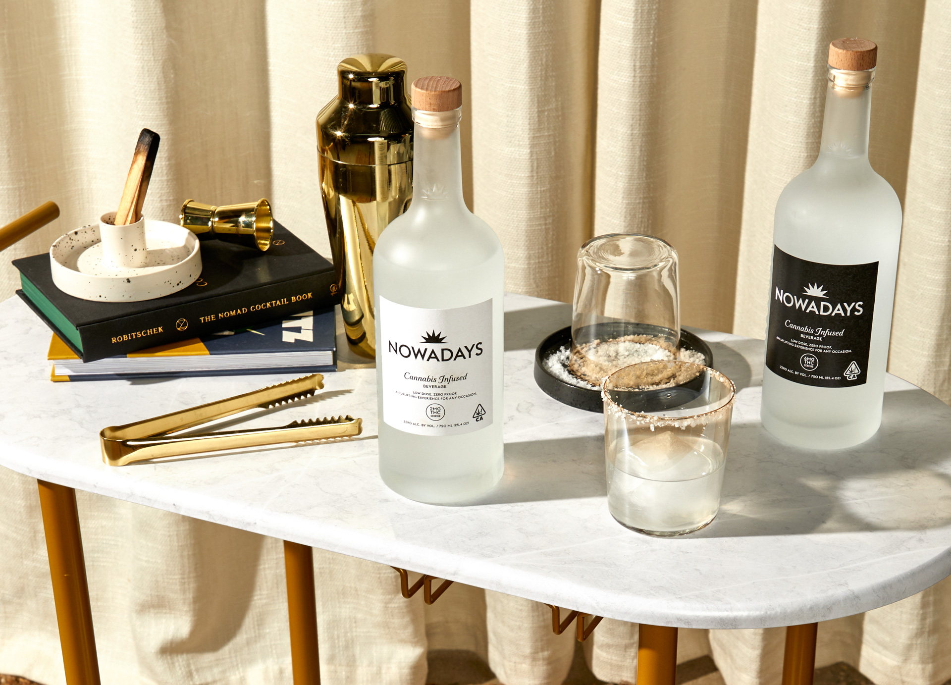 A white barcart with a bottle of Nowadays, cocktail glasses, a gold shaker, tongs and books