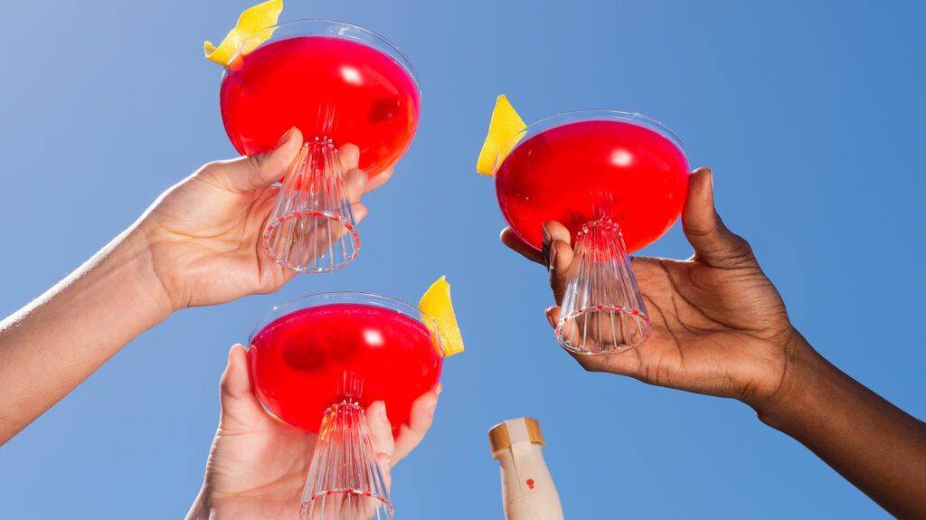 Three hands raising coupe cocktail glasses with a red drink in them to a blue sky backdrop