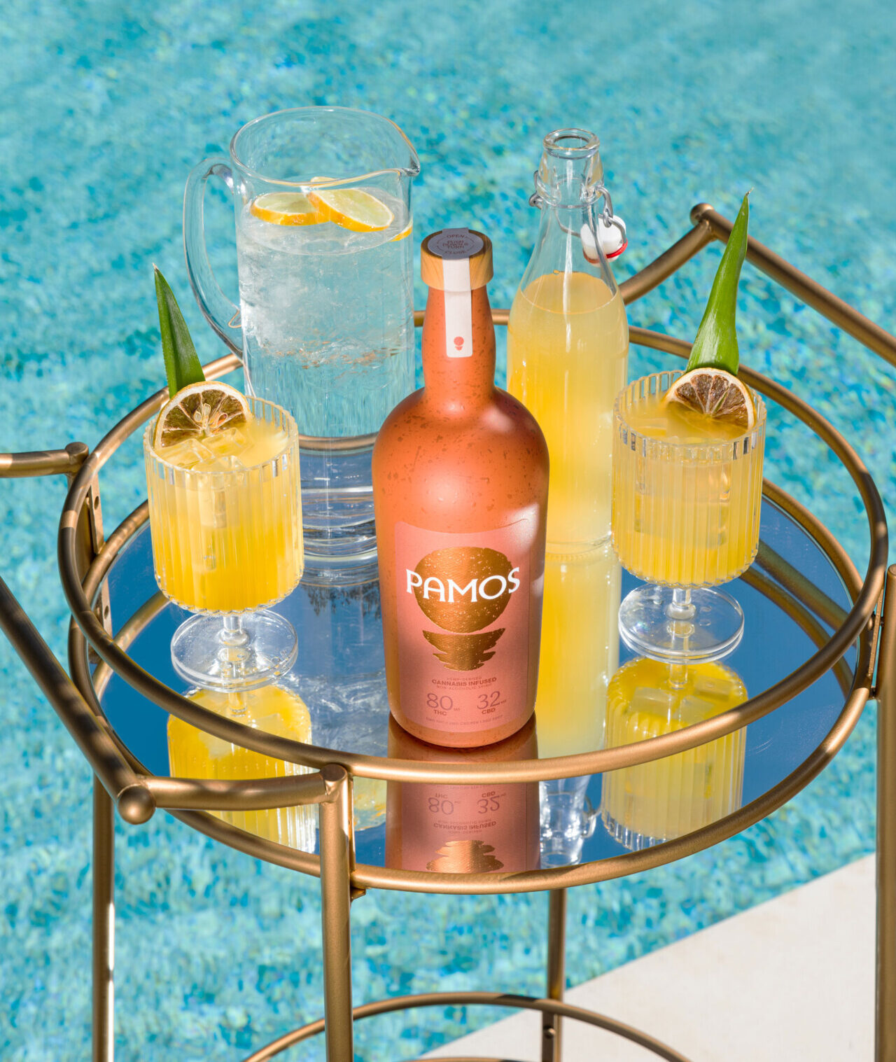A cocktail table by a pool with glasses filled with a yellow cocktail and a bottle of Pamos Spirit on it
