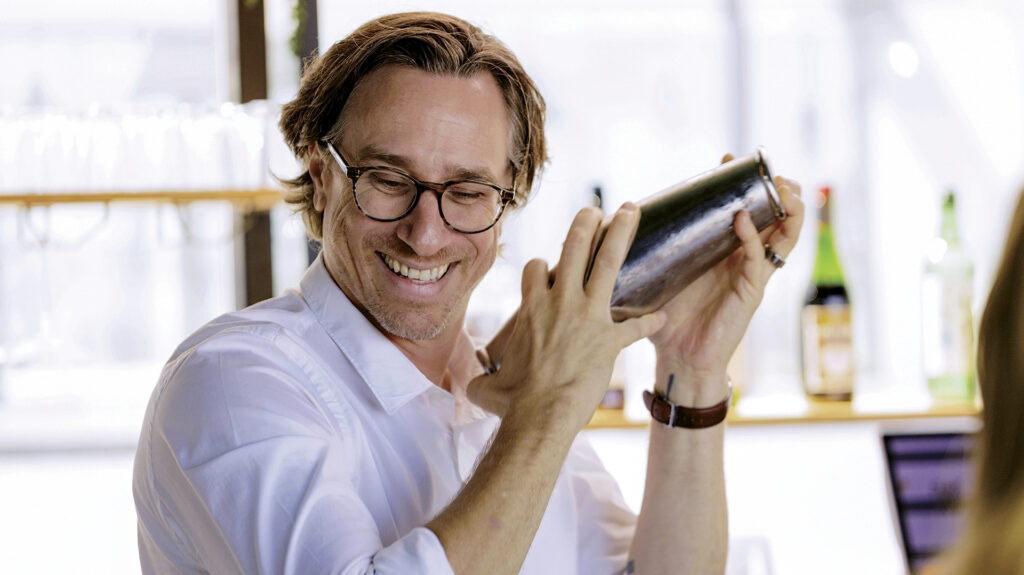 Photo of mixologist Derek Brown, smiling and shaking a cocktail tin in hand
