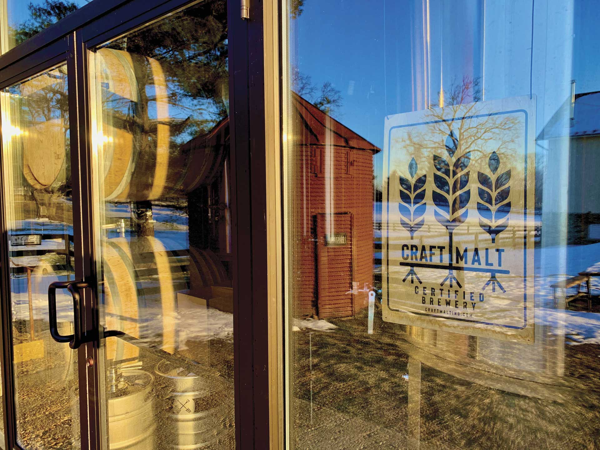 A glass door and window from the outside looking in to see wooden beer barrels and a Craft Malt Certified sign.