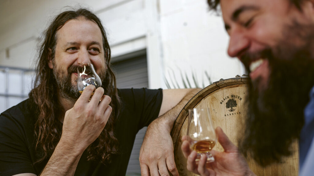 Close up shot of Chris Bower and Rett Murphy of Eda Rhyne Distilling, smiling in front of an oak barrel with tasting glasses in hand of brown liquid.