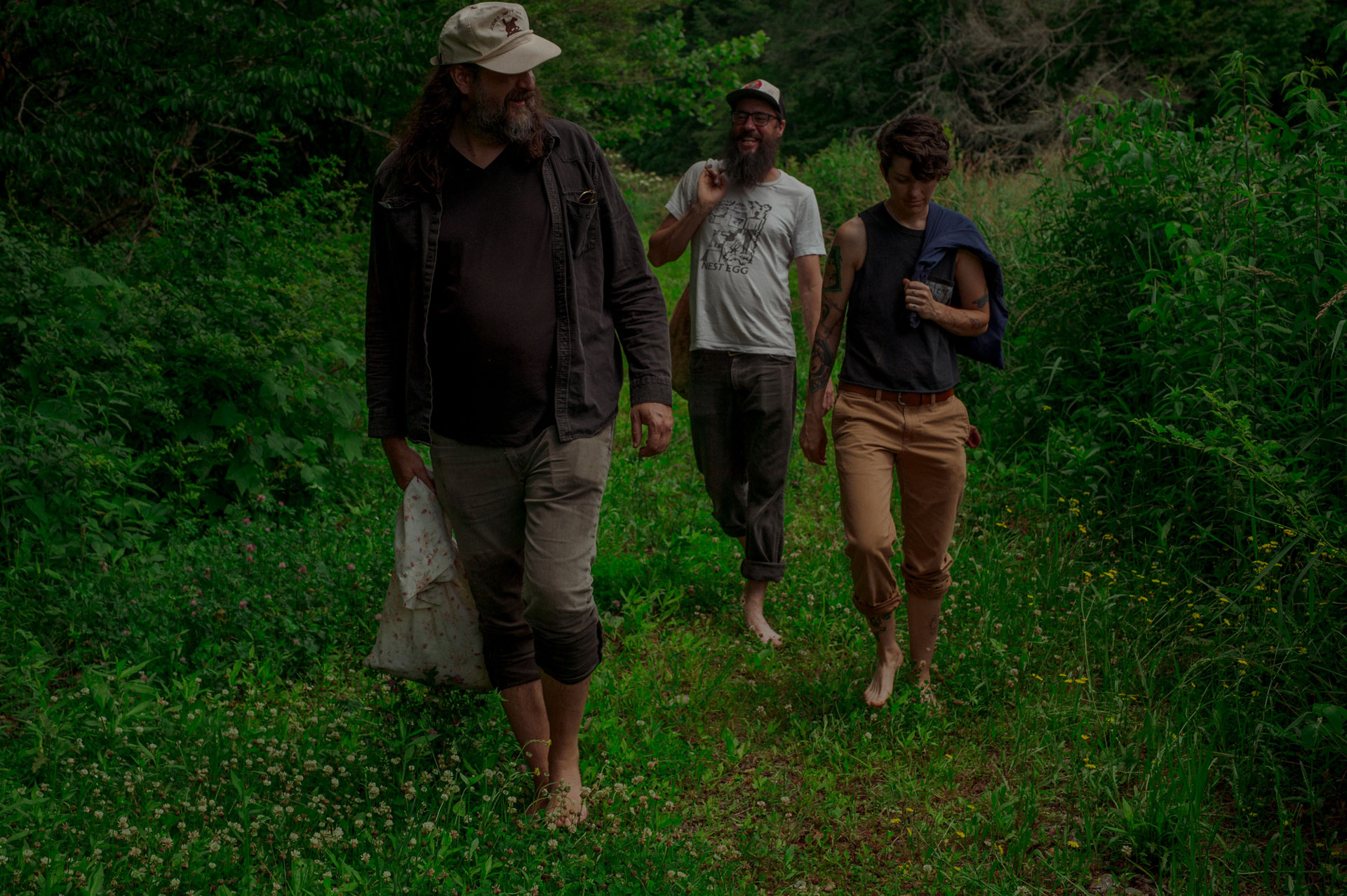 Eda Rhyne Lead Distiller Rider Burton, Rett Murphy and Chris Bower walking barefoot outside to forage herbs, with burlap sacks in hand