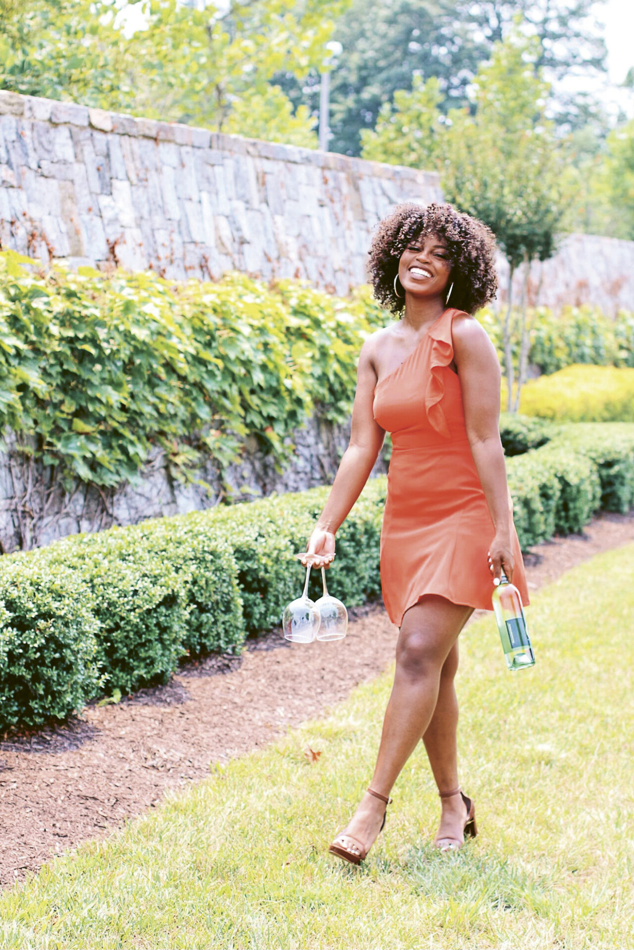 Photograph of Isis Daniel, walking on grass in a lush garden space, holding a wine glass in one hand and wine bottle in the other.