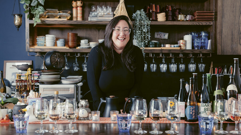 QA-Hero Alyssa DiPasquale smiles from behind the bar with glasses and sake bottles atop in front of her at The Koji Club